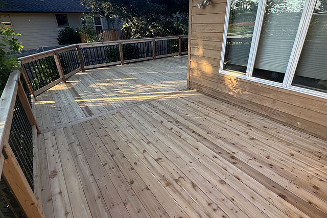 Wide new deck of knotty cedar-toned boards with a board seam across the field, wood post-and-rail railing with black metal balusters, along the back of a tan lap-sided house with large white windows