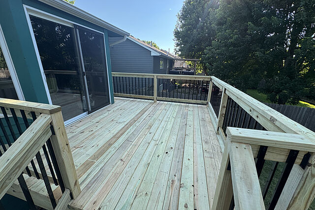 New pale wood deck with wood rails and black metal balusters off a sliding glass door on a teal-sided house, trees beyond