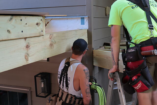 Two workers in branded shirts and tool belts at the back of a house, one steadying an aluminum ladder, with a pressure-treated beam and rim board fitted against the lap siding and a strip of metal flashing above.