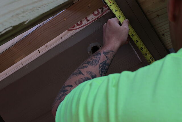 A worker's arm holding a tape measure against brown lap siding beside a strip of flashing tape and exposed sheathing at the edge of a house wall.