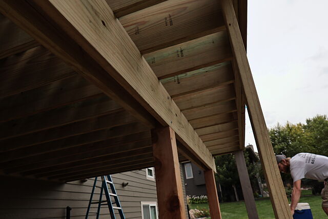 Looking up at the underside of a new pressure-treated deck frame: joists hung on metal hangers off a beam, posts below, a blue ladder against a gray lap-sided house, and a crew member at a cooler on the lawn.