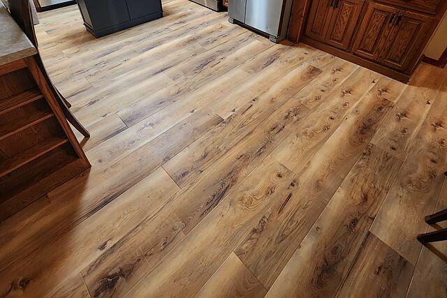 New wide-plank flooring running through a kitchen, tight to oak cabinets, a black island base, and stainless appliances