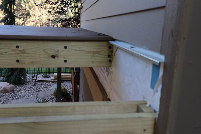 Metal flashing lapped over a treated deck ledger against the house wall, house wrap above