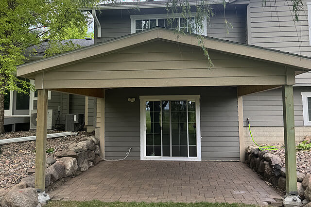 Straight-on view of a front-gable patio cover with a tan lap-sided gable and two treated-wood corner posts on metal standoffs, a shingled roof, over a brick-paver patio at the sliding door of a gray lap-sided two-story house