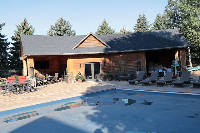 Cedar-sided pool house with a gable entry, French doors, a covered patio with seating and a wall-mounted television, lounge chairs along a covered pool in the foreground, and spruce trees behind.