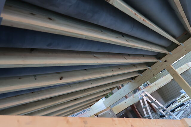 Looking up at covered porch roof framing — evenly spaced rafters running to a beam with a metal tie at each connection, house wrap on the wall behind