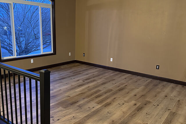 Empty upper-level room with wide-plank wood-look flooring, dark baseboard, a black square-baluster railing with a dark newel post at the top of a stairwell, tan walls, and an arched window at dusk