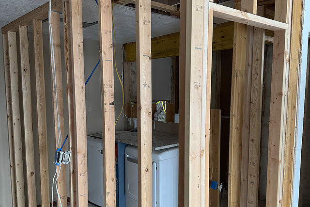 New stud walls with a headered door opening framing in a laundry area, a white washer and dryer under a stone-look countertop, electrical boxes and cable on the studs, the ceiling opened to a beam, and a bare plywood subfloor