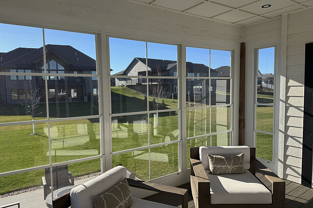 Inside a furnished sunroom on a sunny day: white-framed windows split into stacked panels on three sides, a cedar corner post, a white panel ceiling with a recessed light, a dark plank floor, and lounge chairs